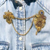 Recto Broche duo Steampunk composée d'engrenages et d'éléments divers (dont une grande feuille) en alliage de bronze reliées par une double chaîne et ornées de petites perles dorées et d'un cristal bleu véritable. portée sur une chemise en jean bleu clair