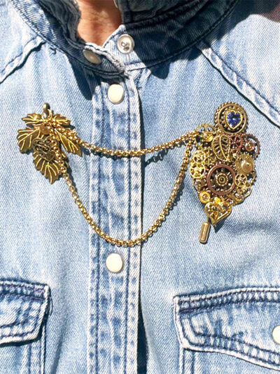 Recto Broche duo Steampunk composée d'engrenages et d'éléments divers (dont une grande feuille) en alliage de bronze reliées par une double chaîne et ornées de petites perles dorées et d'un cristal bleu véritable. portée sur une chemise en jean bleu clair