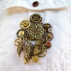 recto Broche Steampunk composée d'un engrenages et d'éléments divers en alliage bronze Ornée de 4 grenats et de petites perles dorées. portée sur la poche d'une chemise blanche