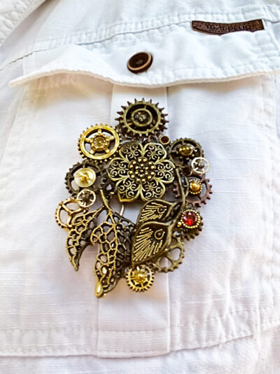 recto Broche Steampunk composée d'un engrenages et d'éléments divers en alliage bronze Ornée de 4 grenats et de petites perles dorées. portée sur la poche d'une chemise blanche