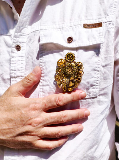 recto Broche Steampunk composée d'un engrenages et d'éléments divers en alliage bronze Ornée de 4 grenats et de petites perles dorées. portée sur la poche d'une chemise blanche avec une main visible